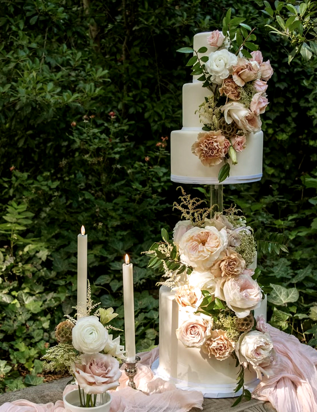 Elegant three-tier wedding cake decorated with white and blush pink flowers, greenery, and gold accents, set outdoors with greenery background and lit candles nearby.