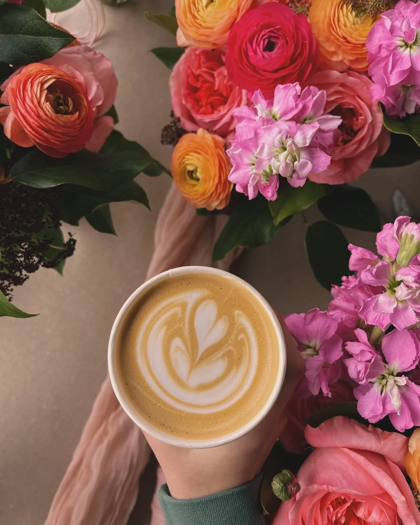 A person holding a cup of latte with foam art in front of a colorful bouquet of pink, orange, and yellow flowers on a beige surface.