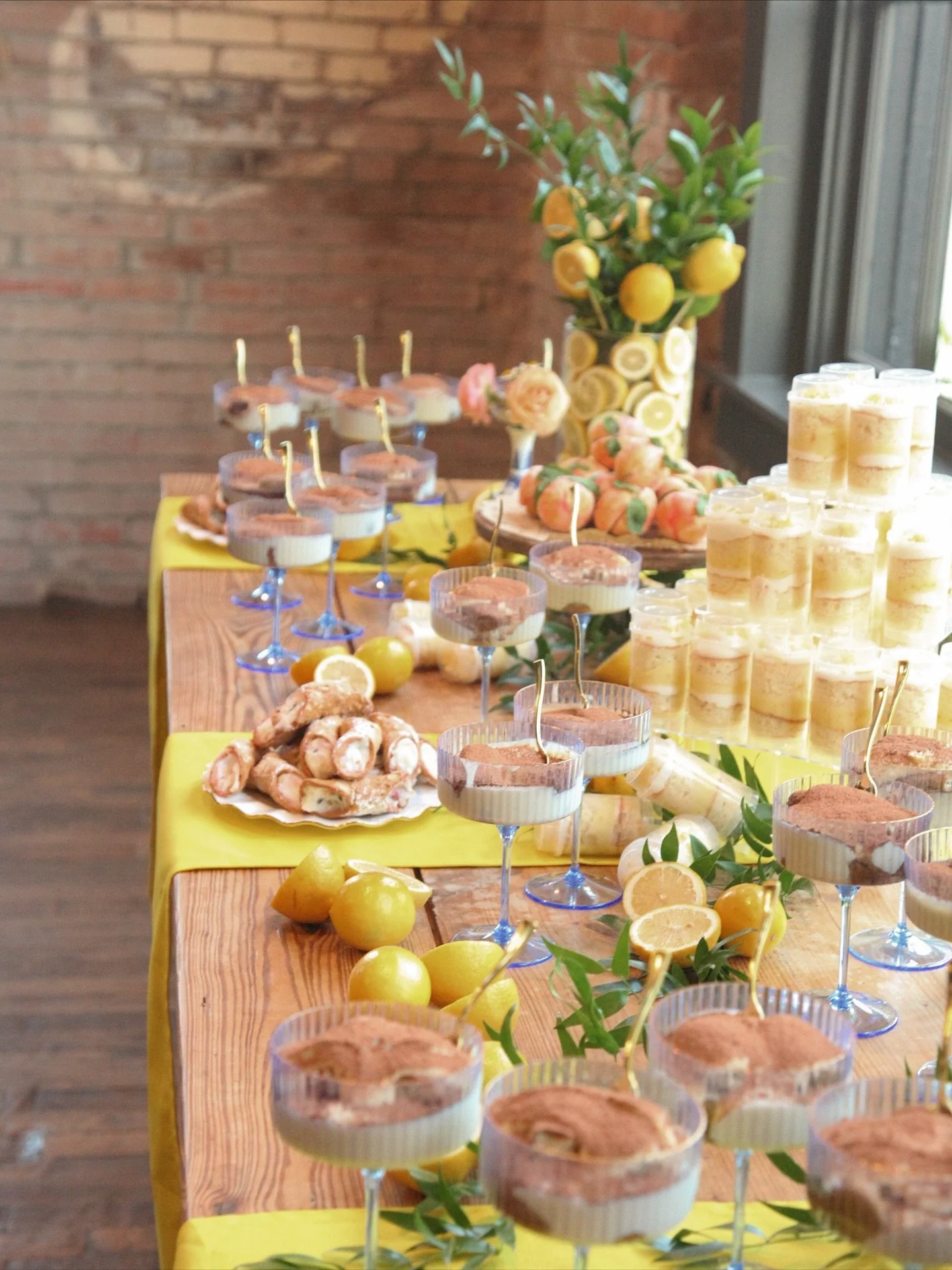 Table with yellow tablecloth holding lemon-themed decorations, desserts including layered mousse cups and croissants, and a large vase of lemons and lemon trees, set against a brick wall.