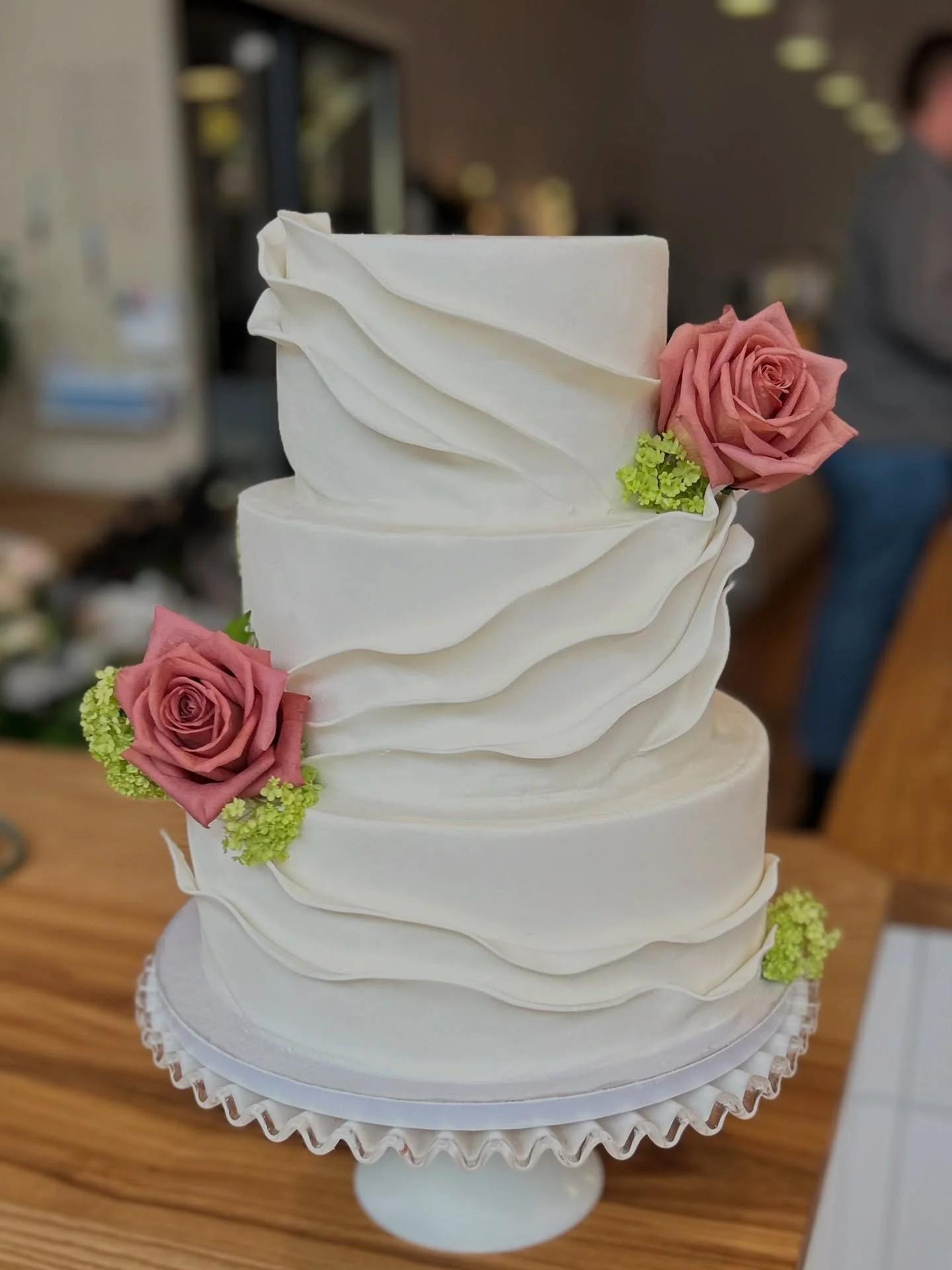 A three-tiered white wedding cake decorated with pink roses and green flowers, placed on a white stand.