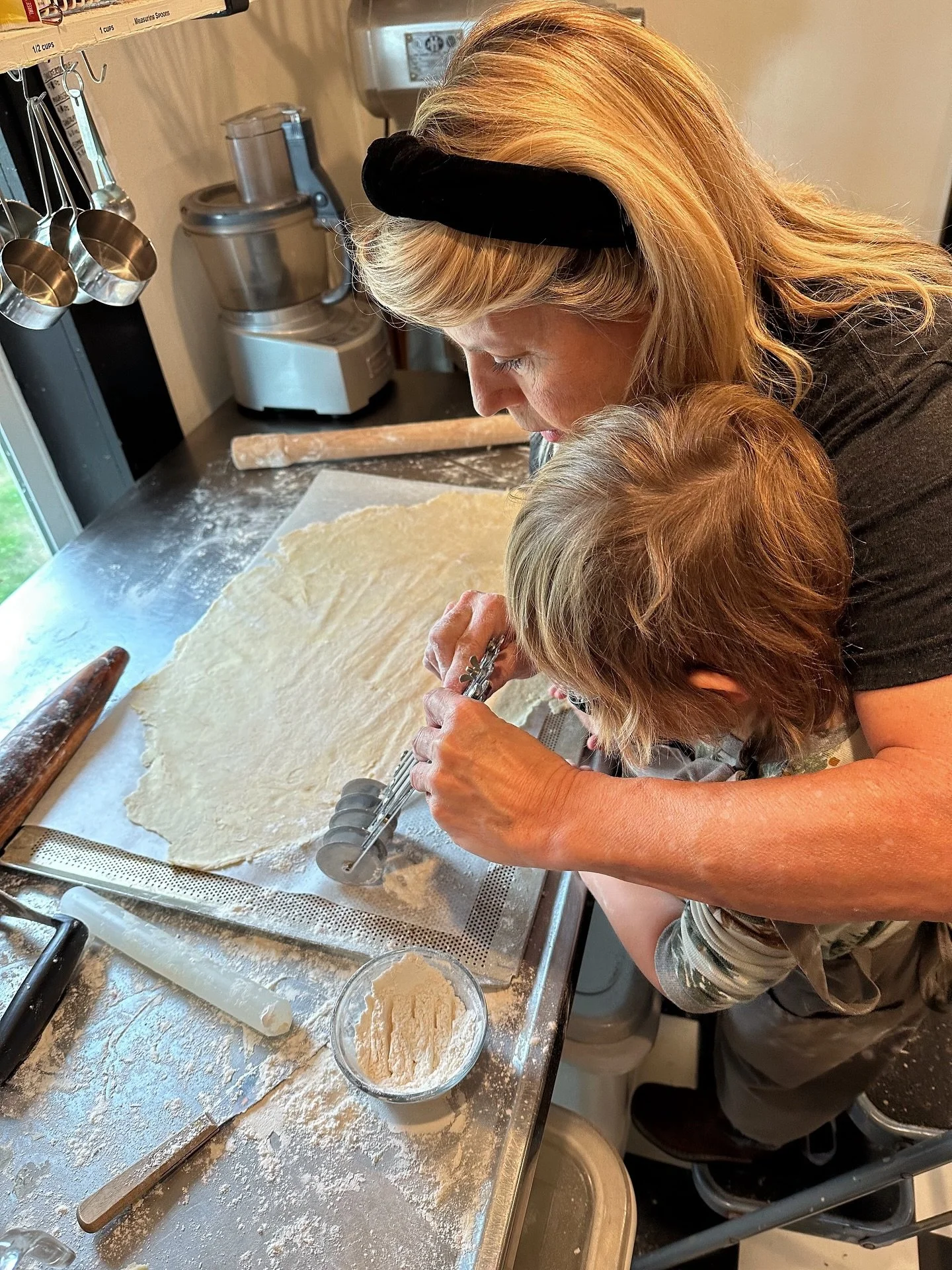 Two women are baking together in a kitchen. One woman is younger and the other older. They are kneeling over a floured surface, using a pastry cutter to cut dough into round shapes. Baking tools and utensils are scattered on the kitchen counter.