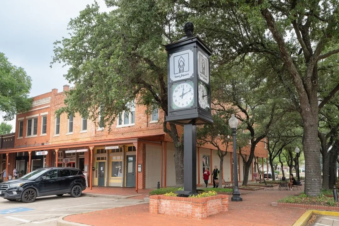 A town square with a clock tower, brick buildings, parked cars, trees, benches, and people walking.
