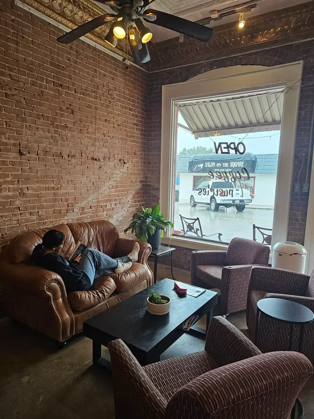 A cozy coffee shop interior with a brown leather couch where a woman is sitting and reading a book. There's a black coffee table with a potted plant, and a patterned armchair nearby. A large window with hanging lettering indicating "OPEN," "COFFEE," and other words allows natural light. Outside, a white van is parked on the street.