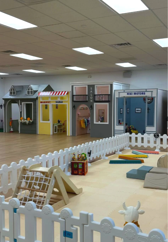 Indoor children's play area with miniature storefronts including a market, salon, and mini-motors, surrounded by a white picket fence and soft play equipment.