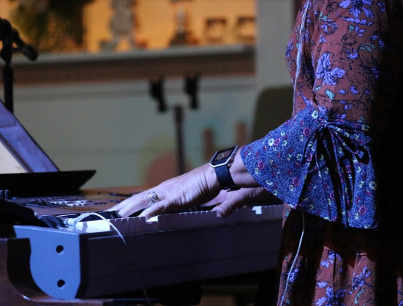 Person playing a digital piano keyboard, wearing a floral-patterned dress, in a room with blurred art and decor in the background.