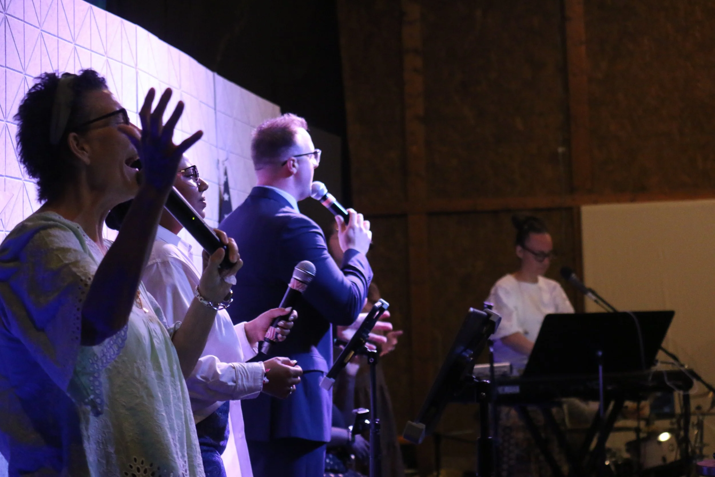 Group of diverse singers performing on stage with microphones, a woman in white dresses and a man in a blue suit, while a woman in the background operates sound equipment.