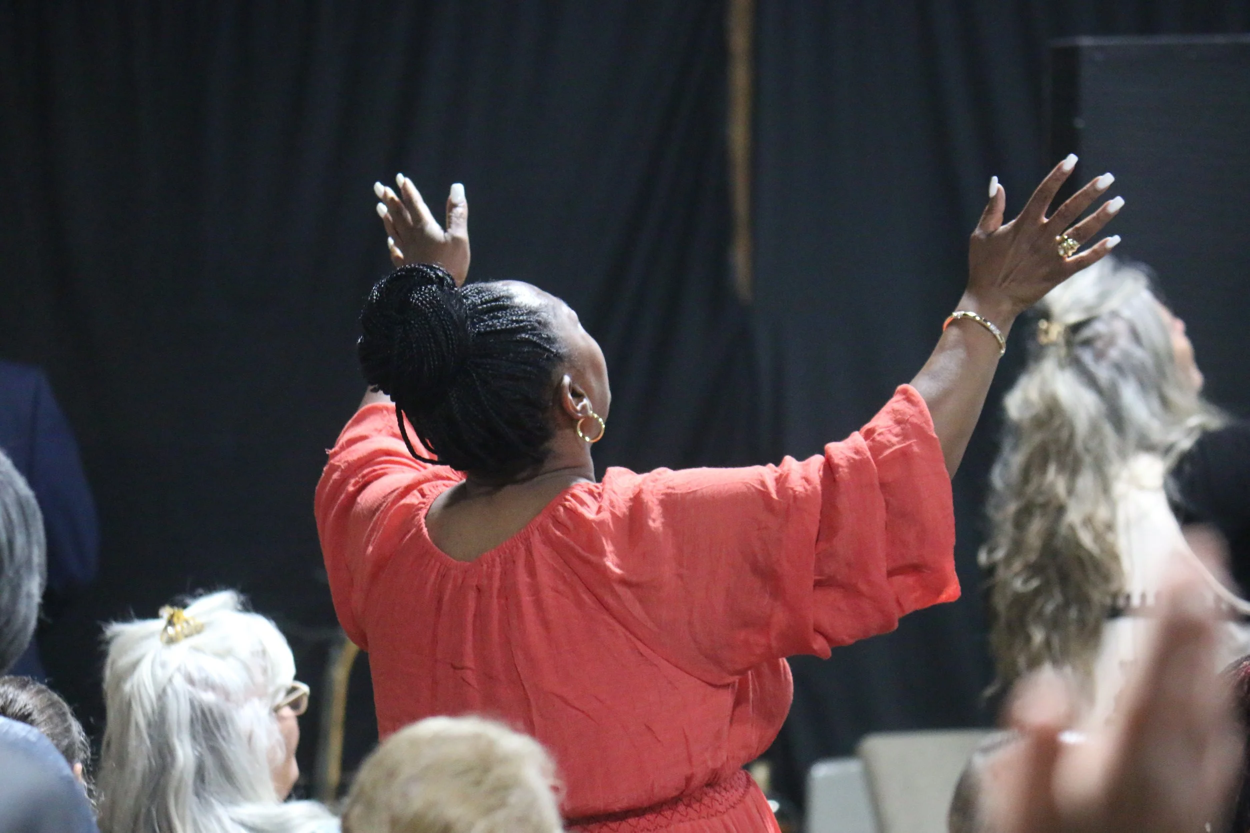 A woman with braided hair tied up in a bun, wearing a coral-colored dress, has her eyes closed and arms raised, seemingly praying or worshiping in a gathering.