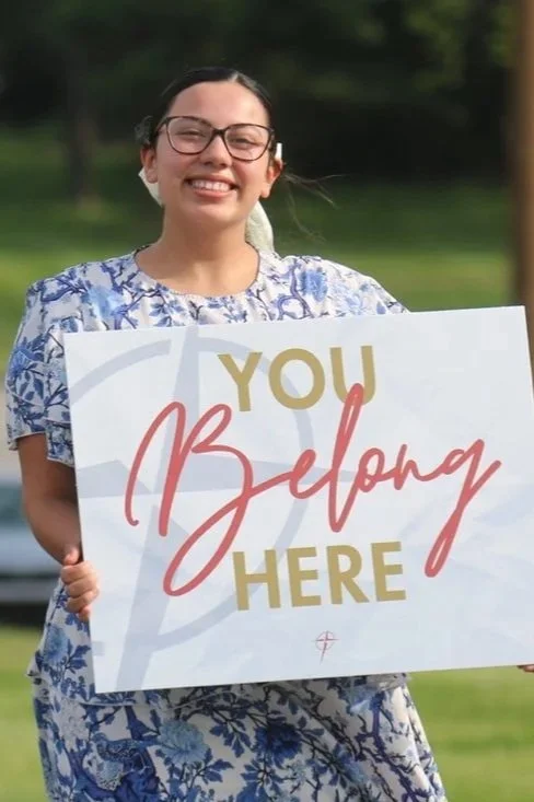 Young woman smiling and holding a sign that reads "You Belong Here" standing outdoors in a park.
