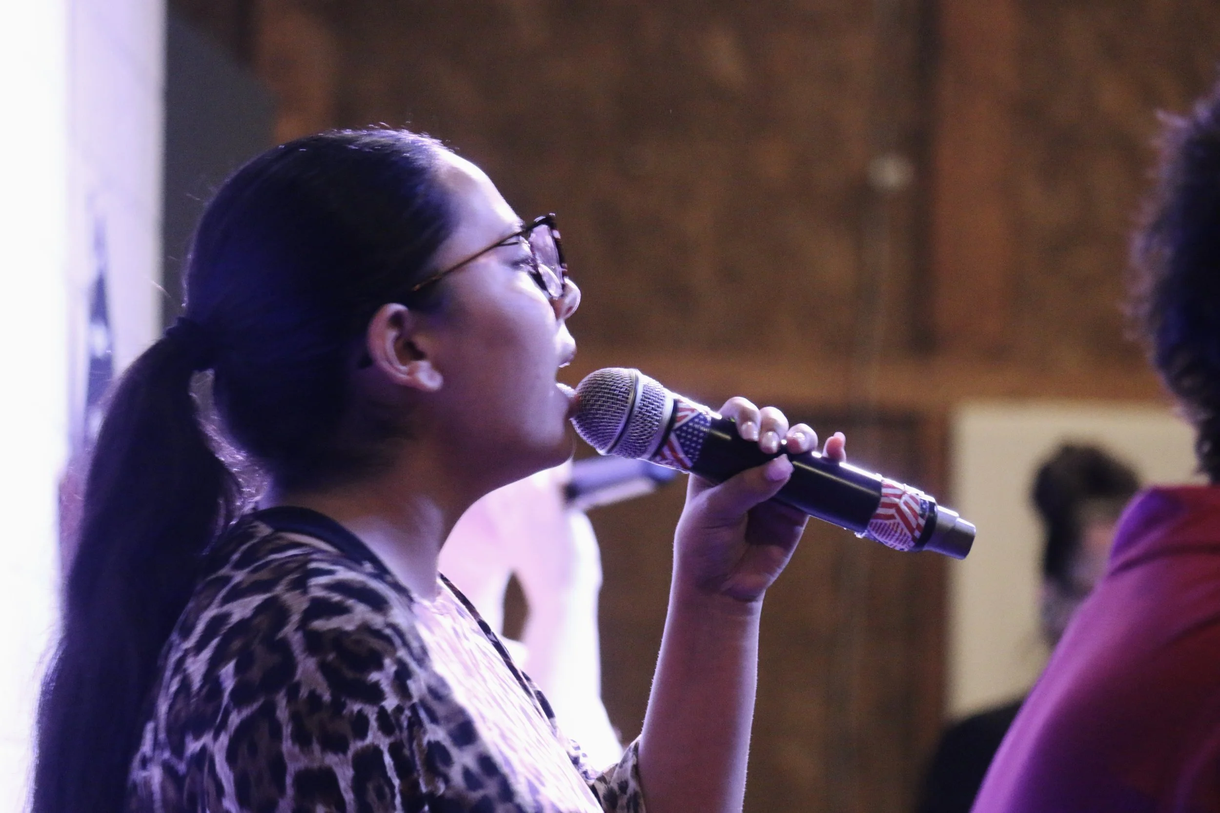 A woman with glasses and a ponytail is speaking into a microphone at an indoor event.