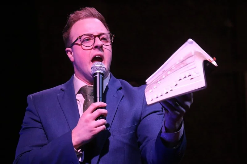 A person in a suit and glasses holding a microphone in one hand and a piece of paper in the other, speaking passionately on stage.