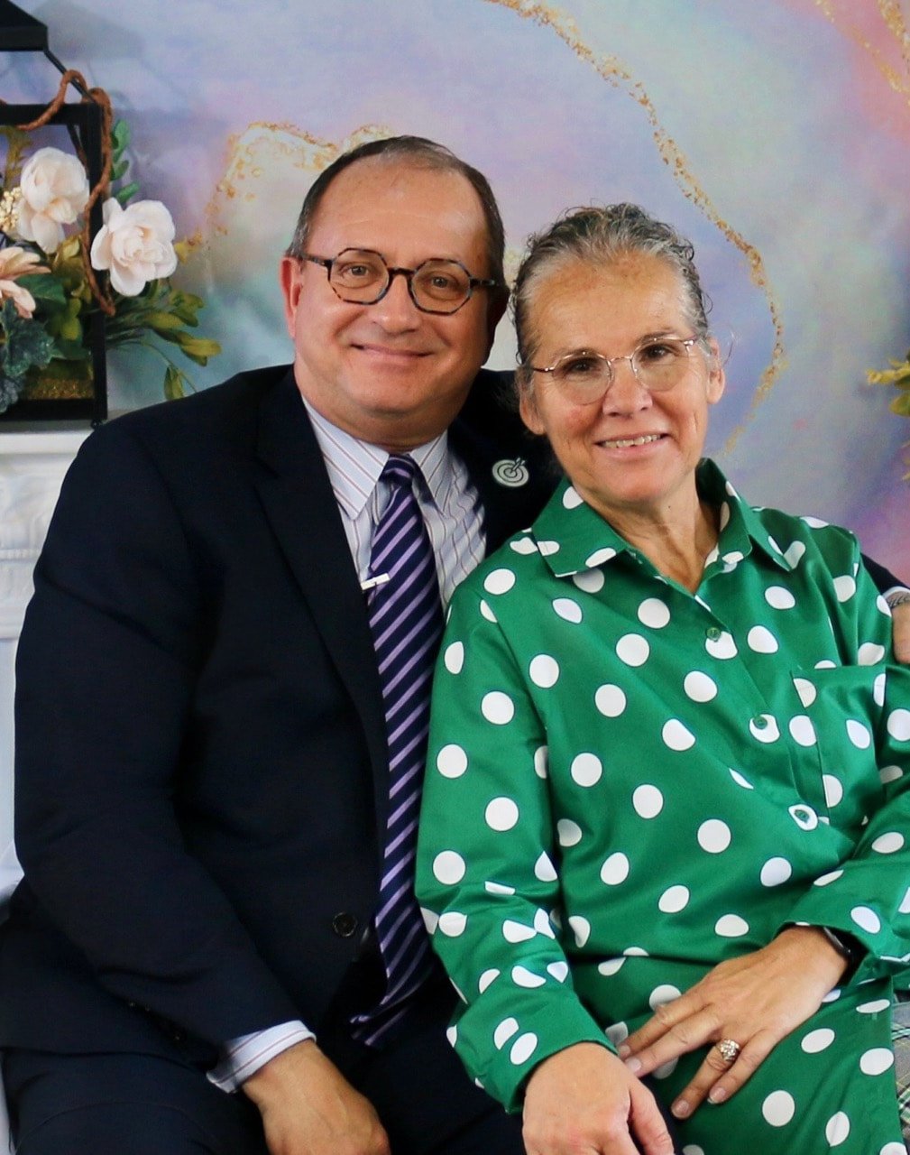 A man and woman sitting together, smiling at the camera, in front of a pastel-colored background with floral decorations.