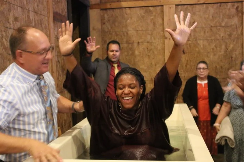 Woman having a baptism experience in a baptismal pool with raised hands, surrounded by smiling and clapping people in a room with wooden walls.
