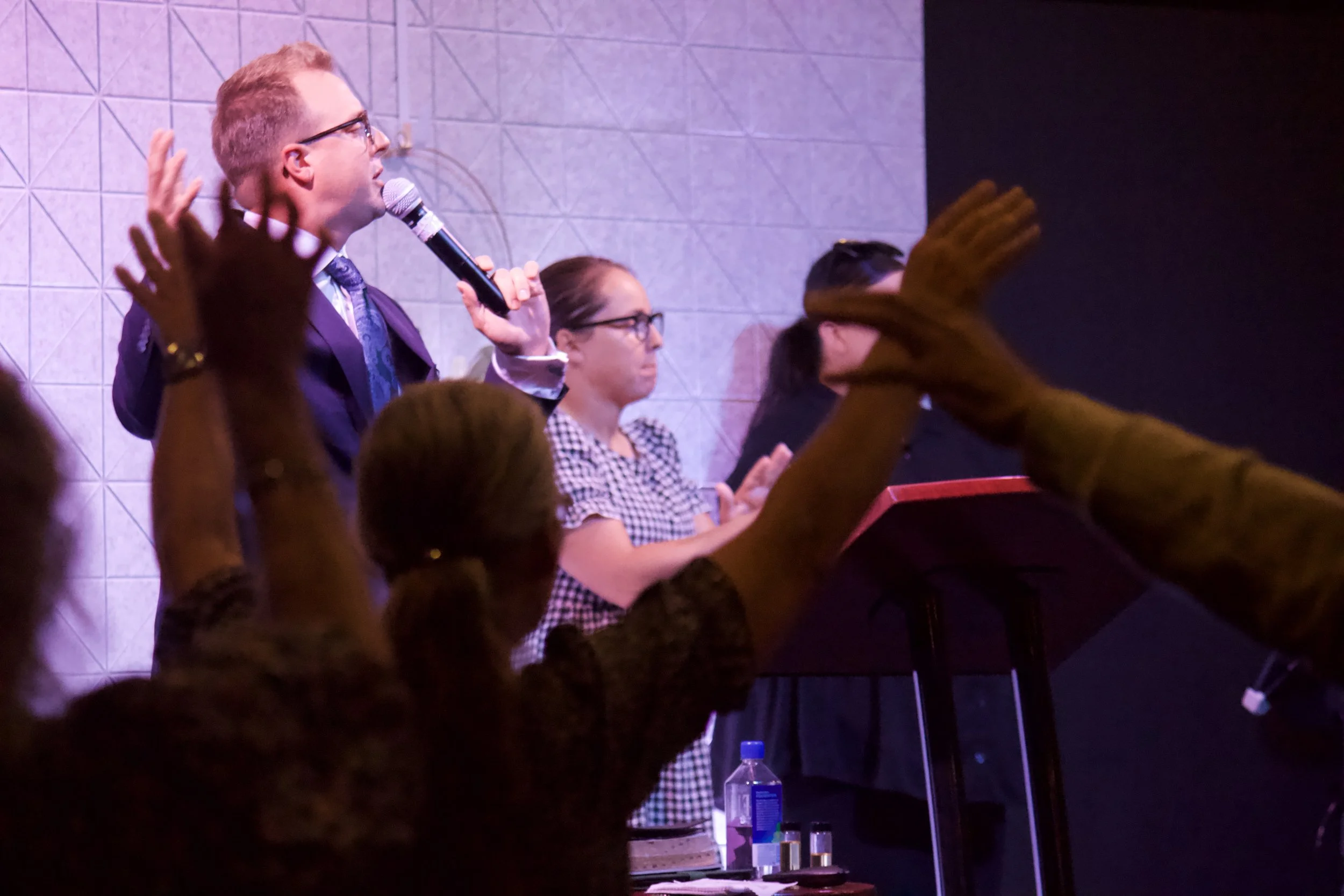 A man with glasses and a suit holding a microphone appears to be speaking or singing during an event. Several audience members are raising their hands, and some are clapping. The background shows a tiled wall and a music stand with a red top.