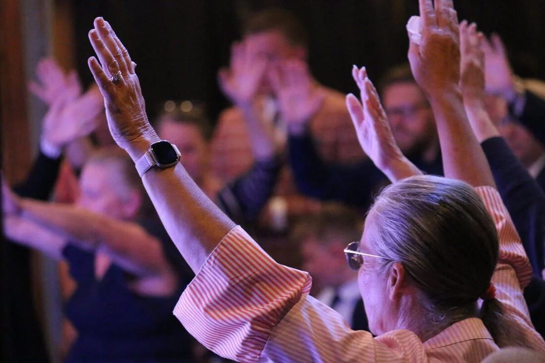A group of people raising their hands in a dark, indoor setting, with a woman in the foreground wearing sunglasses and a striped shirt.