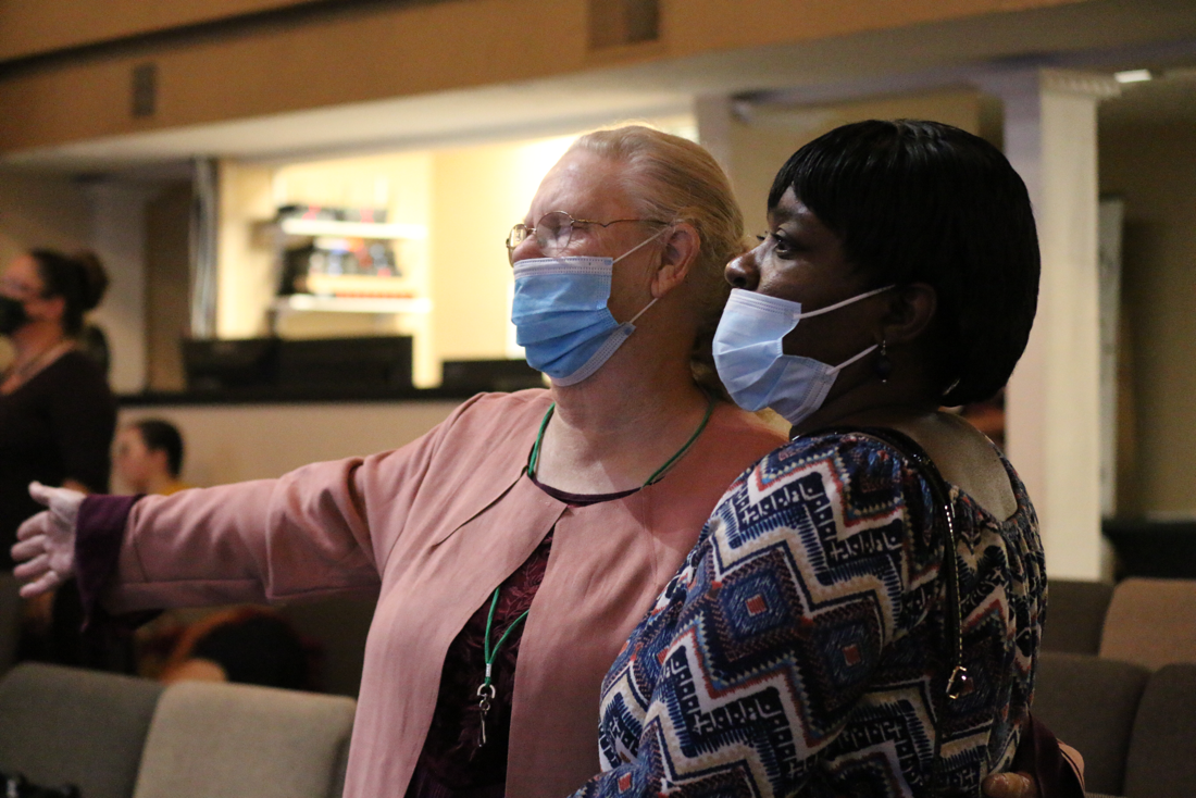 Two women wearing face masks, one with blonde hair in a pink jacket, and the other with short black hair in a patterned dress, are indoors, appearing to be engaged in a conversation or presentation.