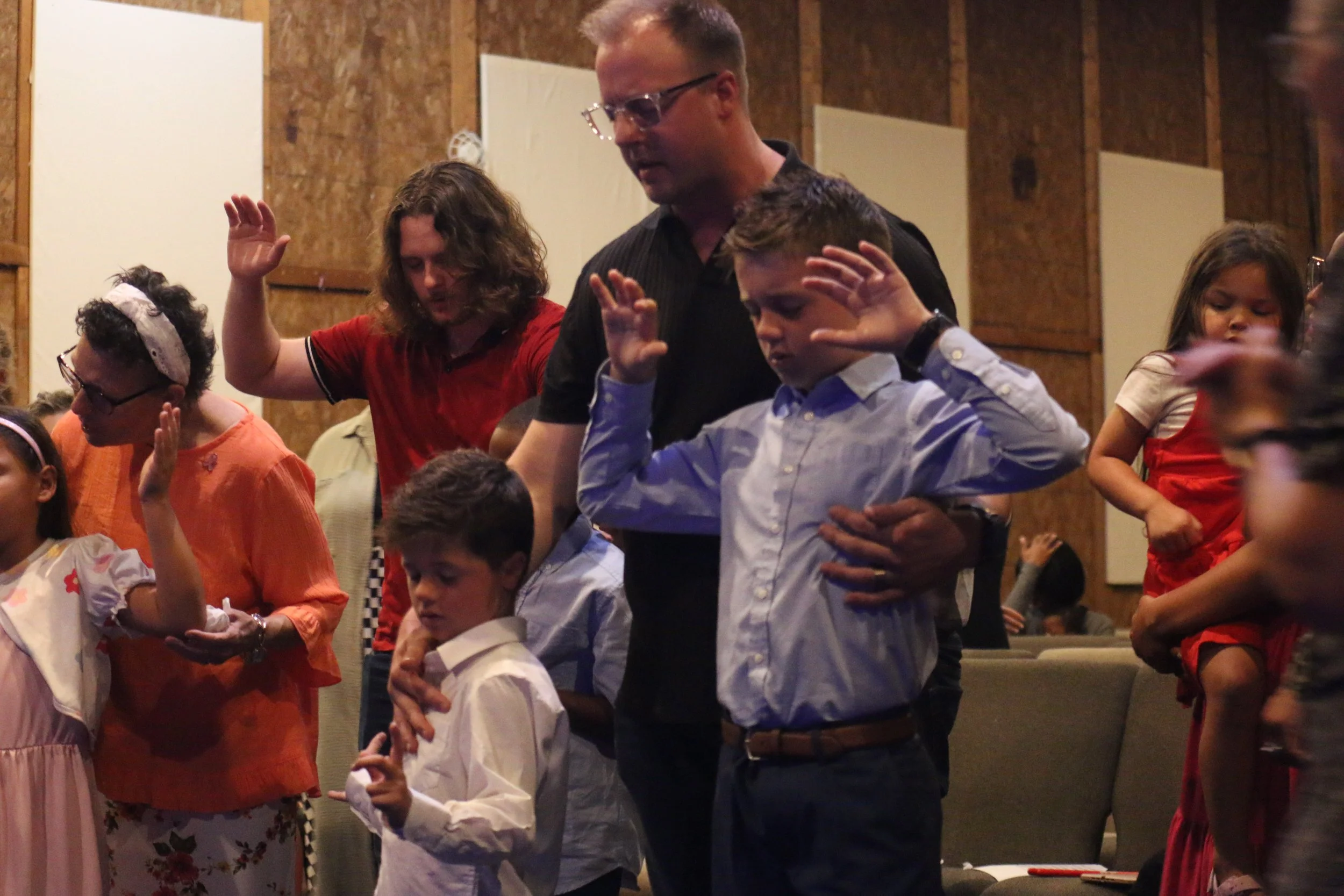 People gathered in an indoor setting, standing with their eyes closed and hands raised, engaged in a prayer or worship activity.