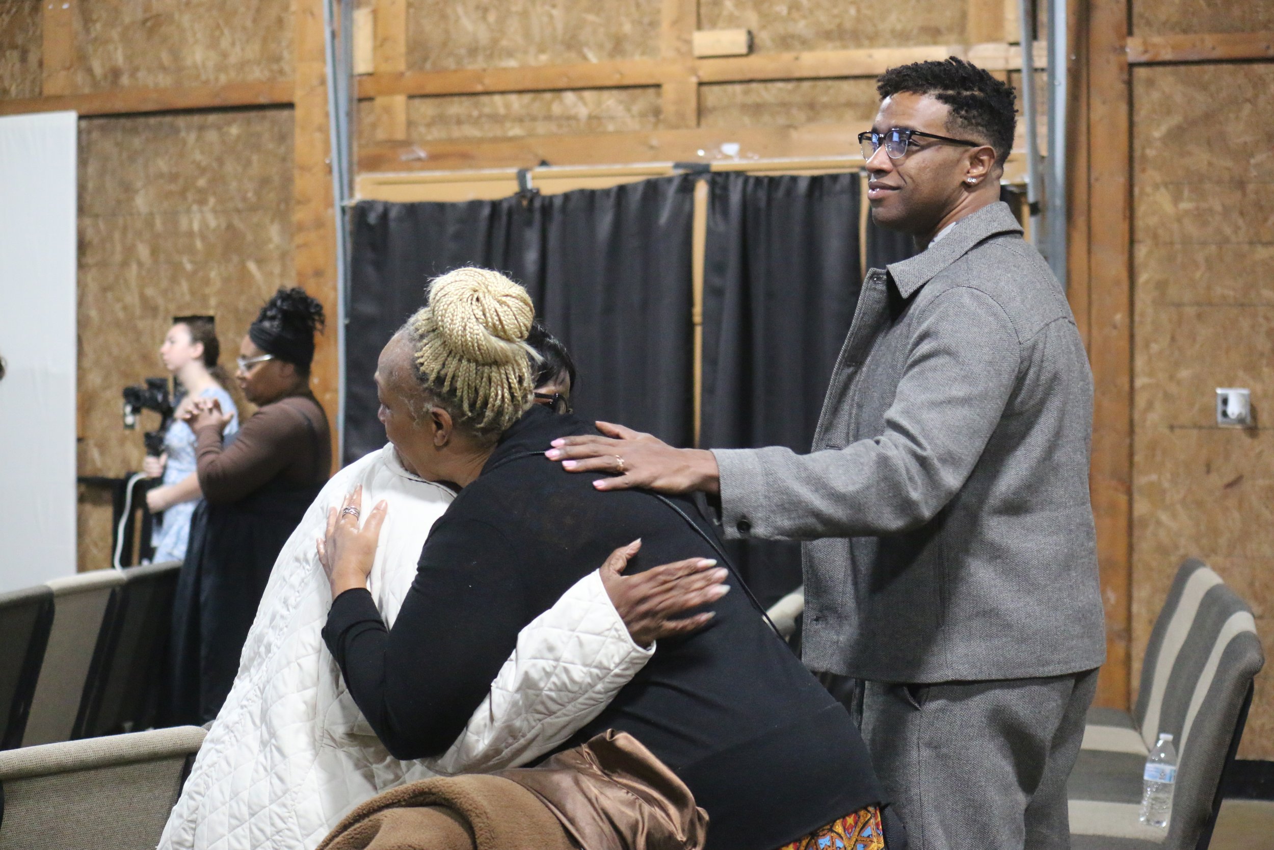 A man gently places his hand on a woman's back as they hug in a church or community hall setting. Several other people are in the background, some observing or involved in activities, with wooden walls and chairs in the space.