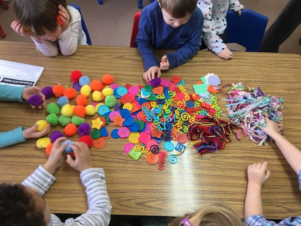Children sitting around a wooden table playing with colorful craft supplies, including pom-poms, buttons, paper shapes, string, and ribbon.