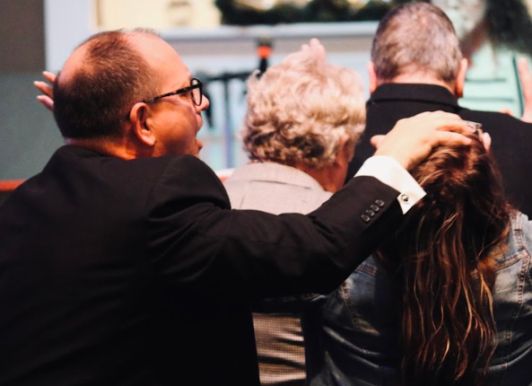 A man in a suit with glasses is gently touching a young woman's head in a crowd, possibly at an outdoor event or gathering.