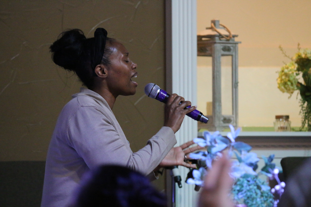 Woman singing or speaking into a microphone during an event or presentation, with floral arrangements in the foreground and a decorative background.