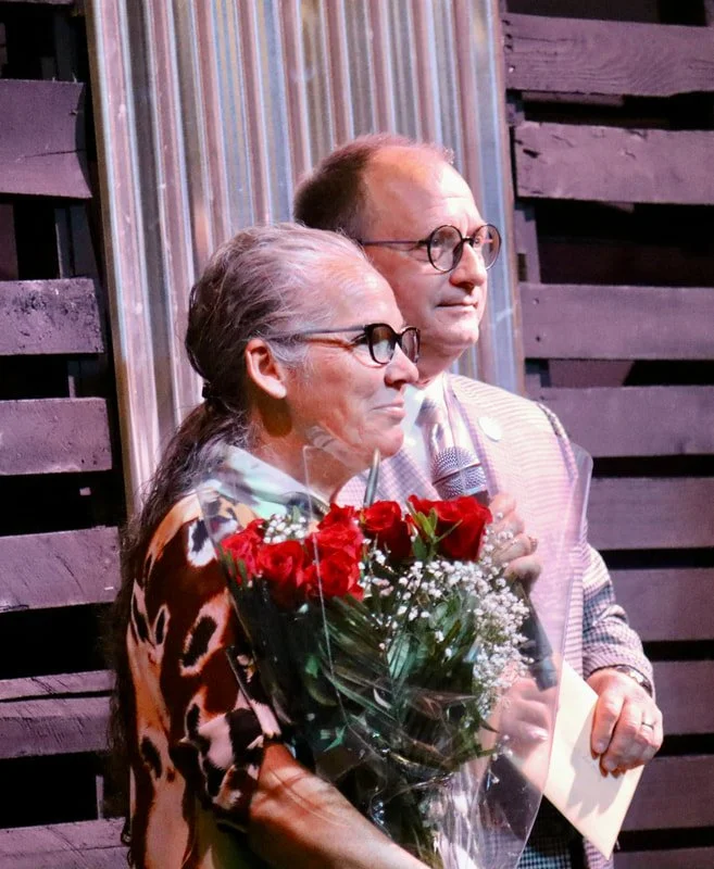 A woman holding a bouquet of red roses and white baby's breath flowers, standing next to a man. They are both wearing glasses and appear to be at an event with a rustic wooden backdrop.