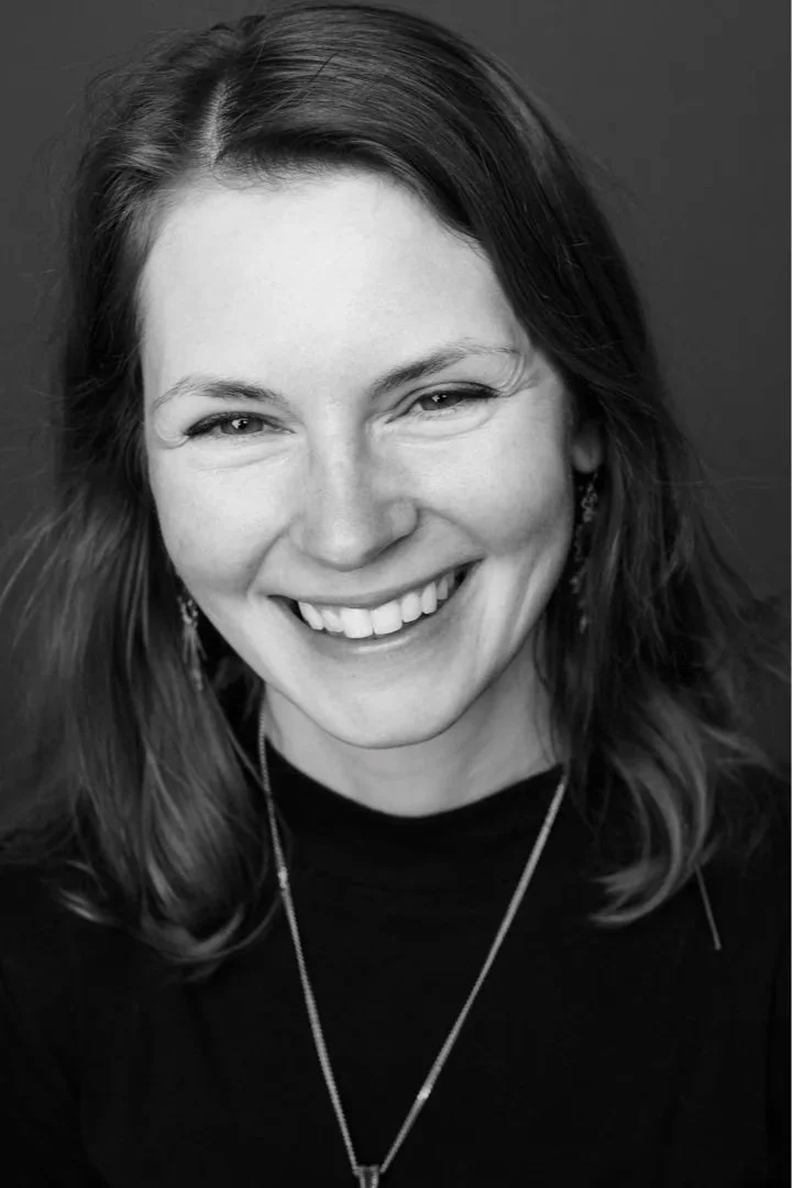 Black and white portrait of a woman with medium-length hair, smiling, with earrings and a necklace, wearing a dark top.