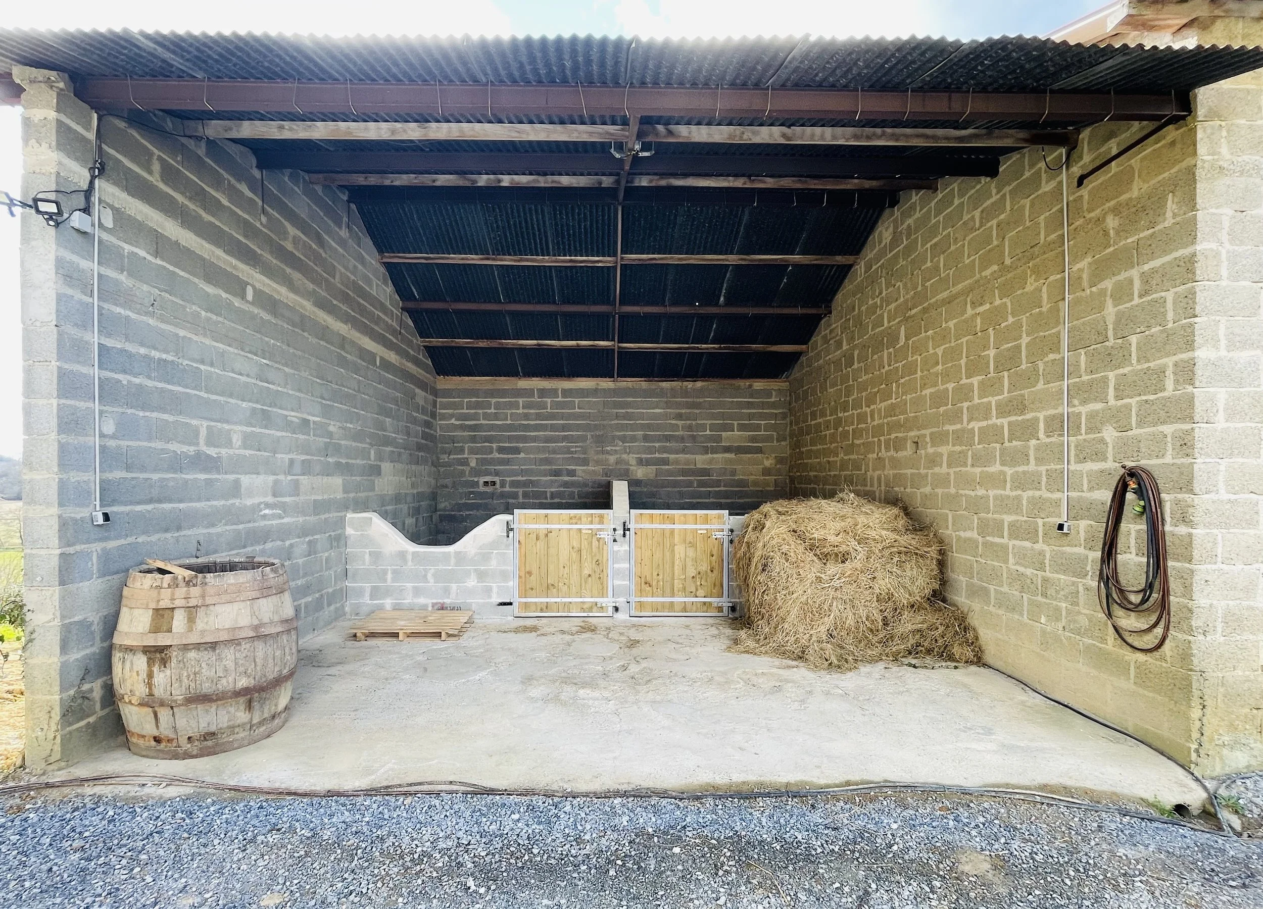 Espace de stockage avec un tonneau en bois, un ouvrage de suture à gauche, deux portes en bois au centre, de la paille en tas à droite, et des câbles électriques accrochés au mur.