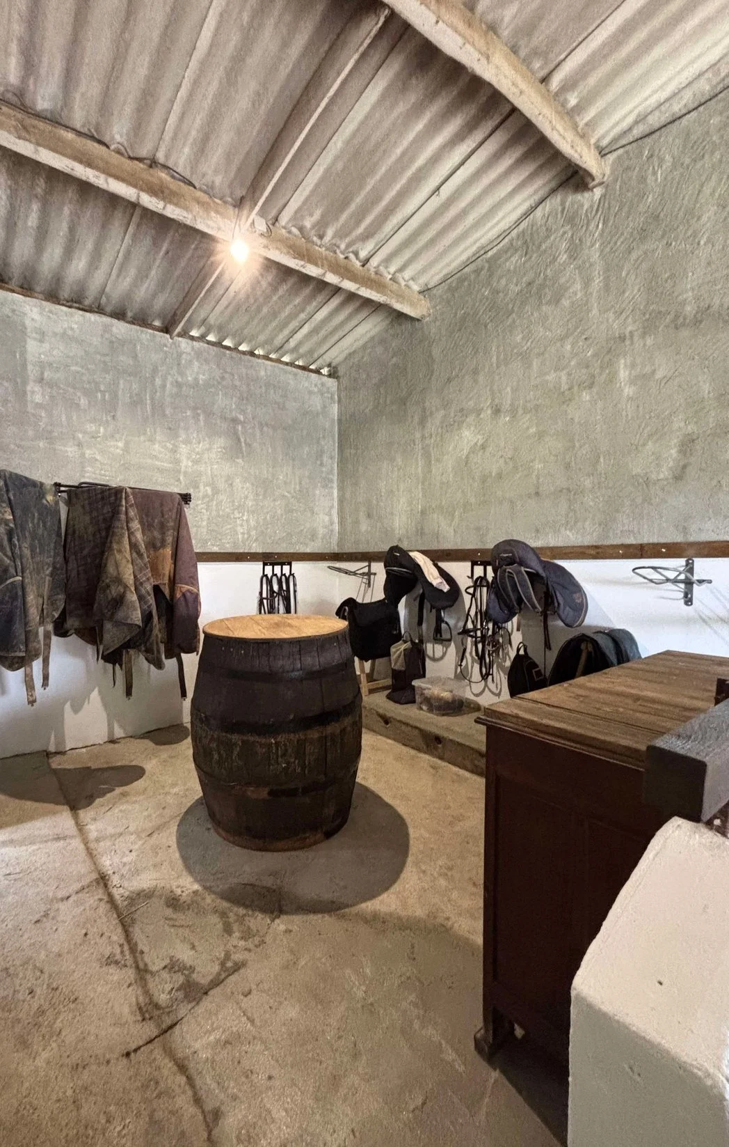 Un espace de rangement ou de sellerie avec des vêtements, casques, et un tonneau en bois dans une pièce avec murs en plâtre et plafond en tôle ondulée.