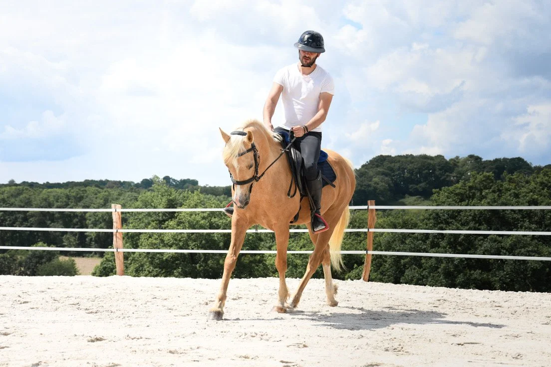 Un homme portant un casque de sécurité monte à cheval dans un enclos extérieur en pleine nature.