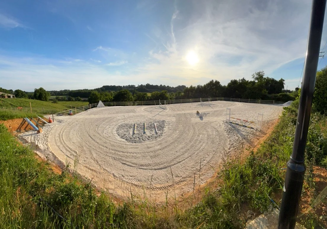Terrain de saut d'obstacles en construction en plein air, avec des obstacles modernes et une clôture en bois, sous un ciel en partie nuageux avec le soleil en position presque haute.