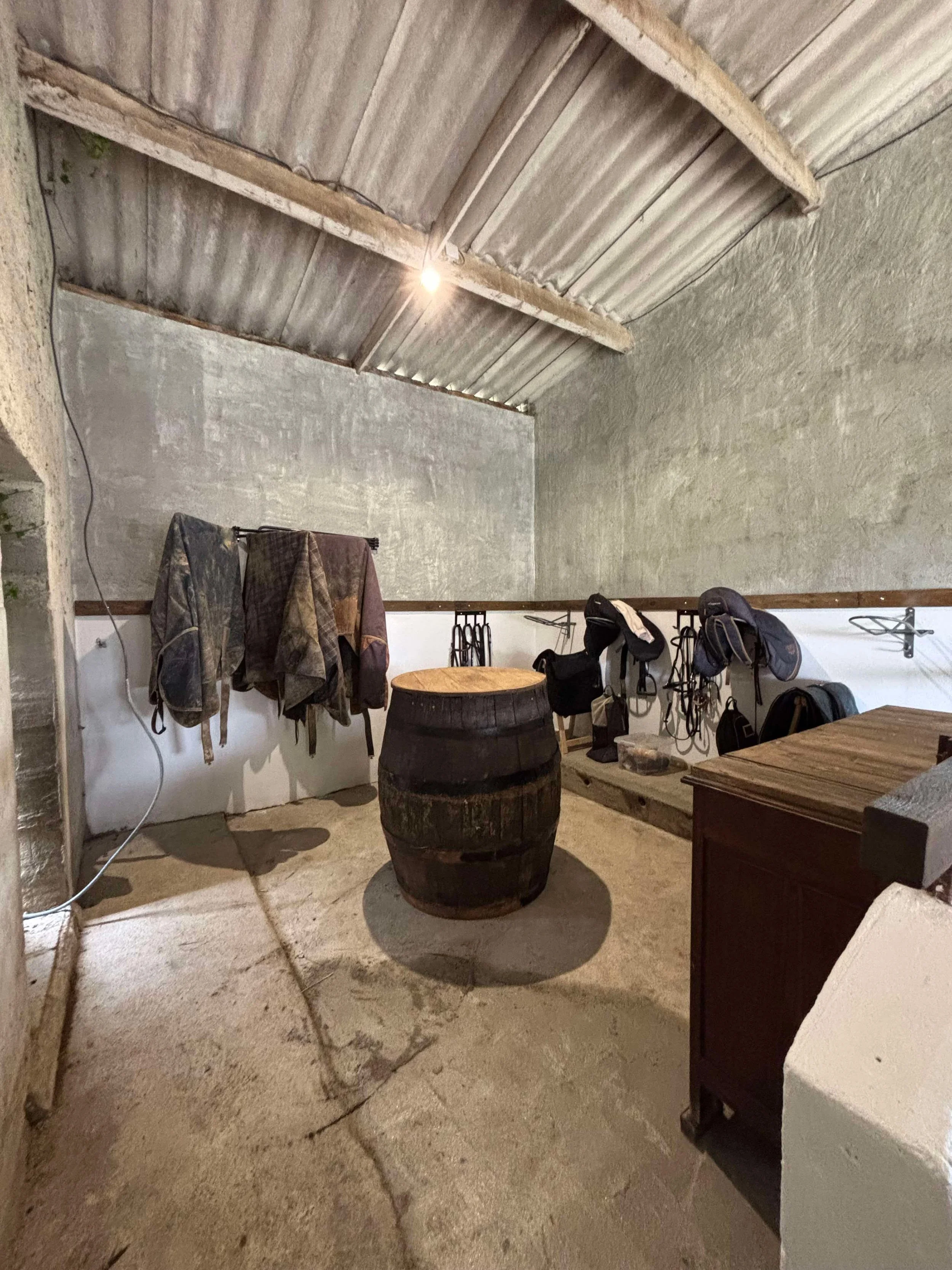 Intérieur d'une pièce rustic avec des vêtements et des sacs suspendus sur une corde, une vieille cher à baril en bois au centre, et un meuble en bois dans un coin.