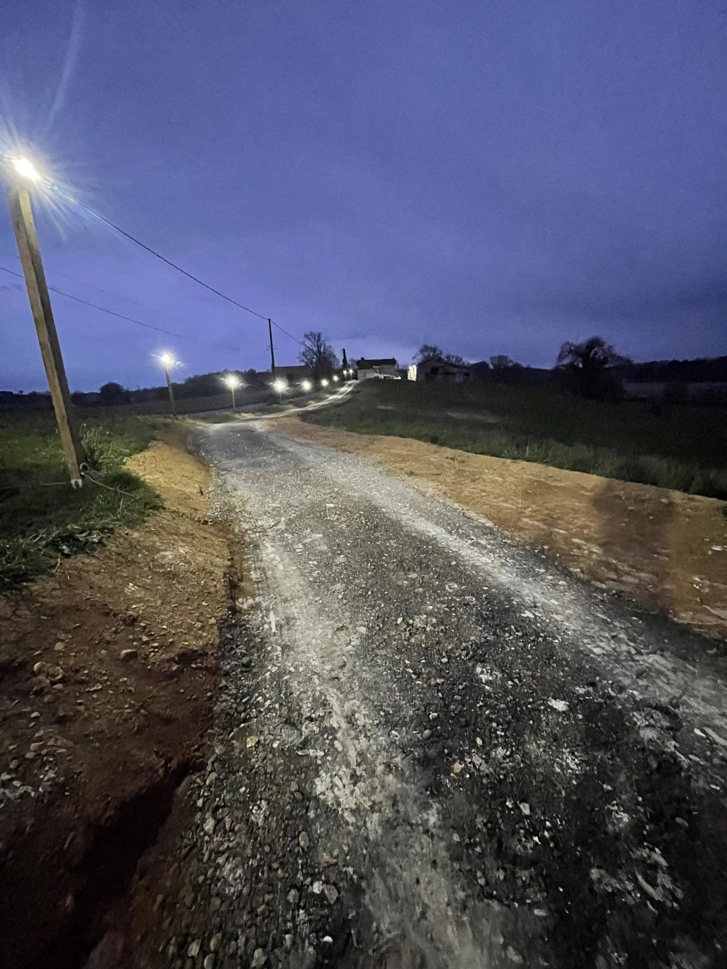 Chemin non asphalté avec des lampadaires le long de la route, en campagne, sous un ciel crépusculaire ou nocturne.