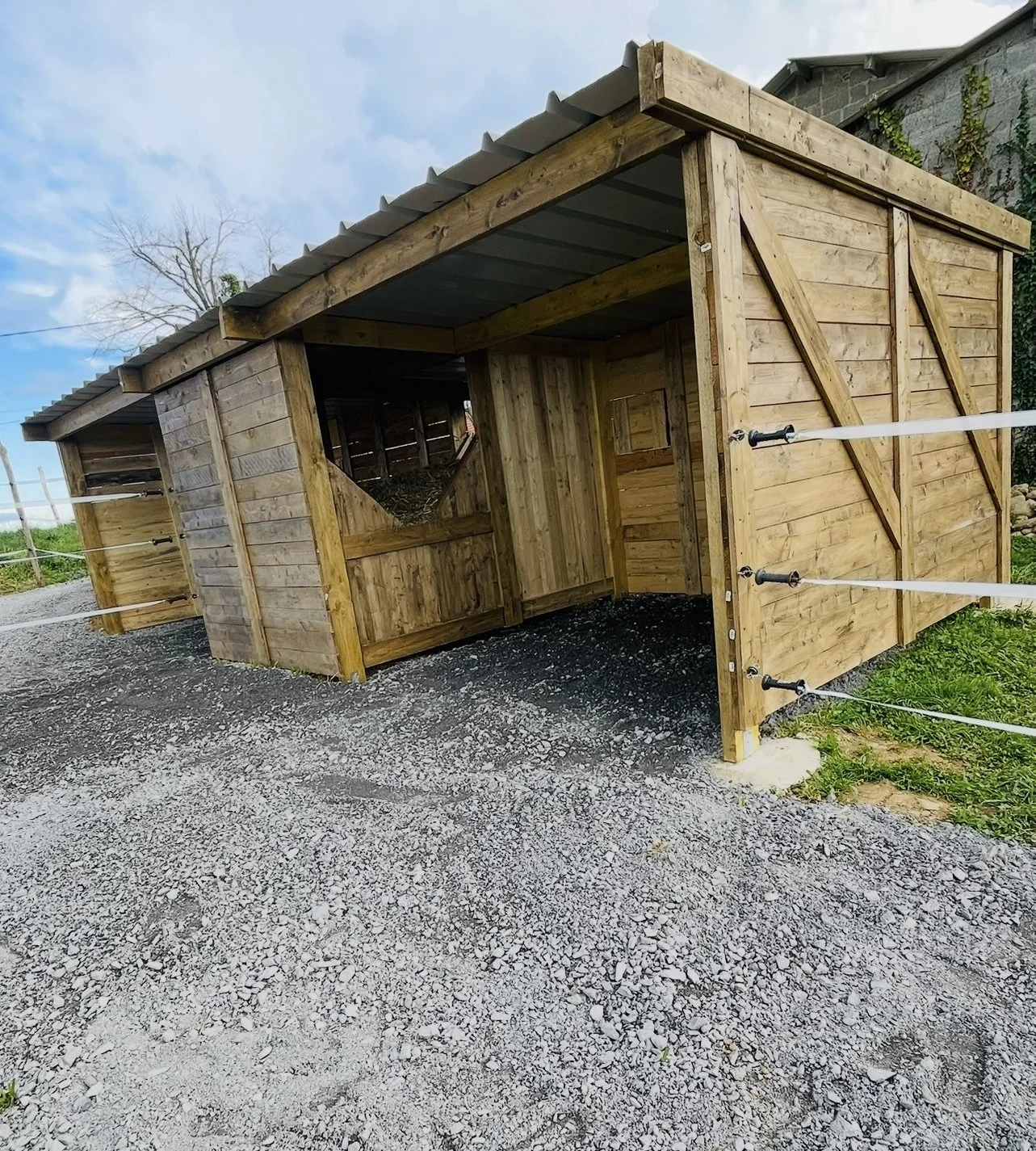 Abri pour animaux en bois avec parois en planches, toit en tôle, et ouvert sur un côté, posée sur un sol gravillonné.