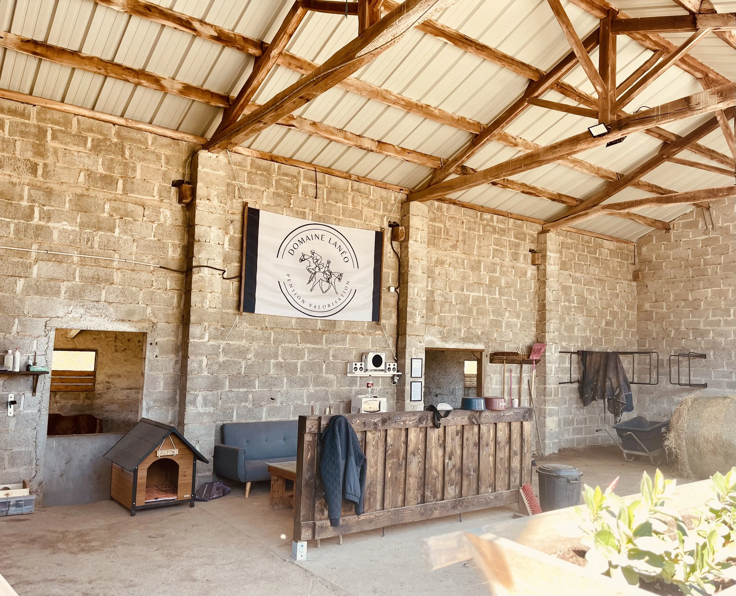Intérieur d'une ferme ou d'un hangar avec murs en briques et toits en bois, contenant un mur avec une bannière, un canapé, un abreuvoir pour animaux, et diverses équipements agricoles.
