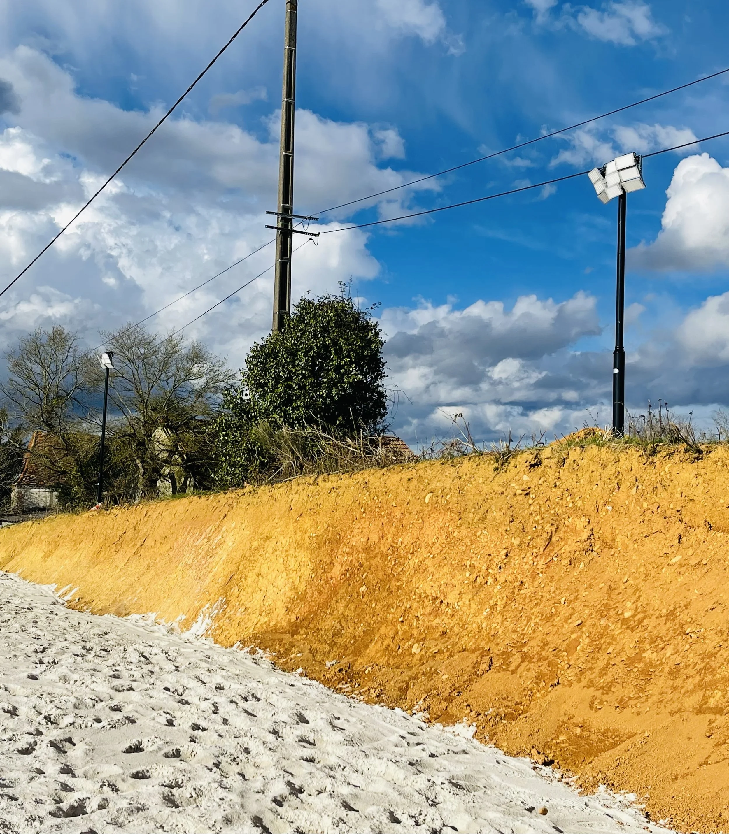 Un chemin en sable devant une digue en terre orange, avec des lampadaires, un poteau électrique, des arbres sans feuilles et un ciel bleu avec des nuages blancs.