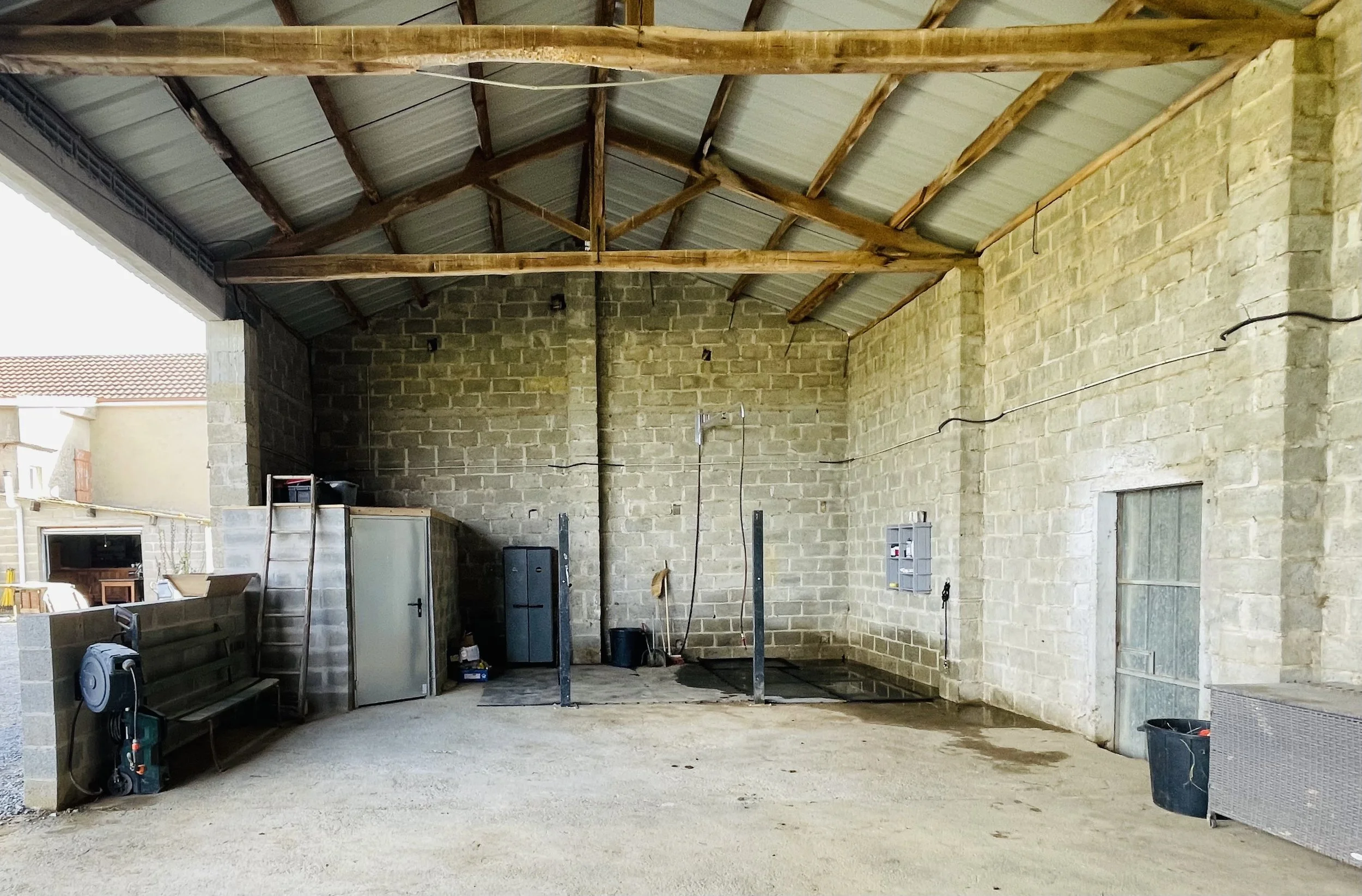 Un grand espace couvert avec murs en pierre, partie du sol en terre battue, un mobilier et des outils de jardinage. Le toit est en métal avec des structures en bois.