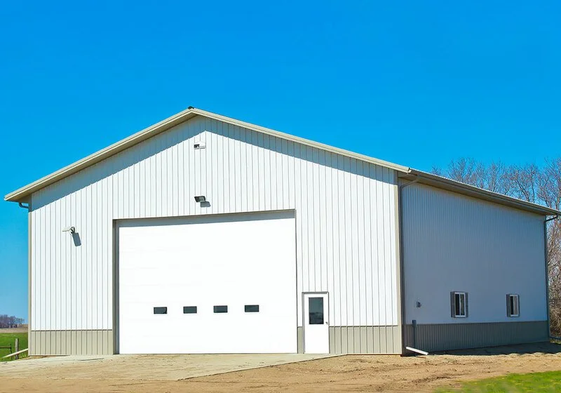 Large white metal building with a roll-up garage door and a standard entry door, set in a rural area under a clear blue sky.