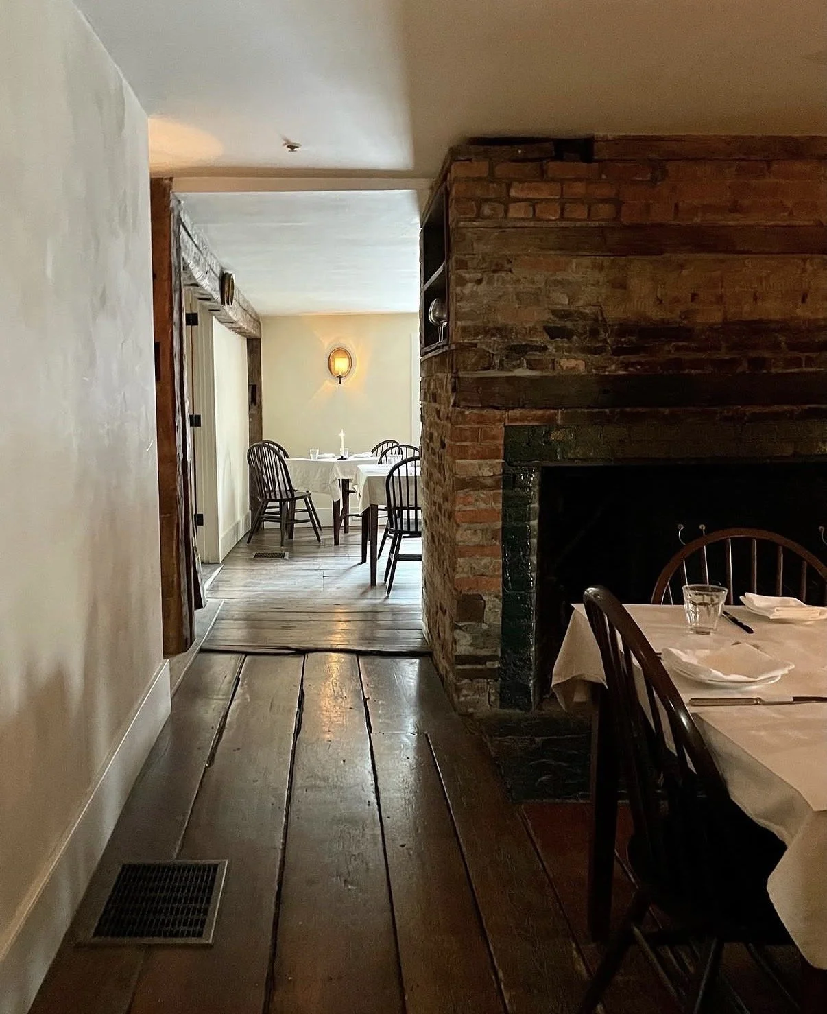 Interior view of a cozy restaurant with wooden floors, a brick fireplace, and wooden chairs around tables set for dining.