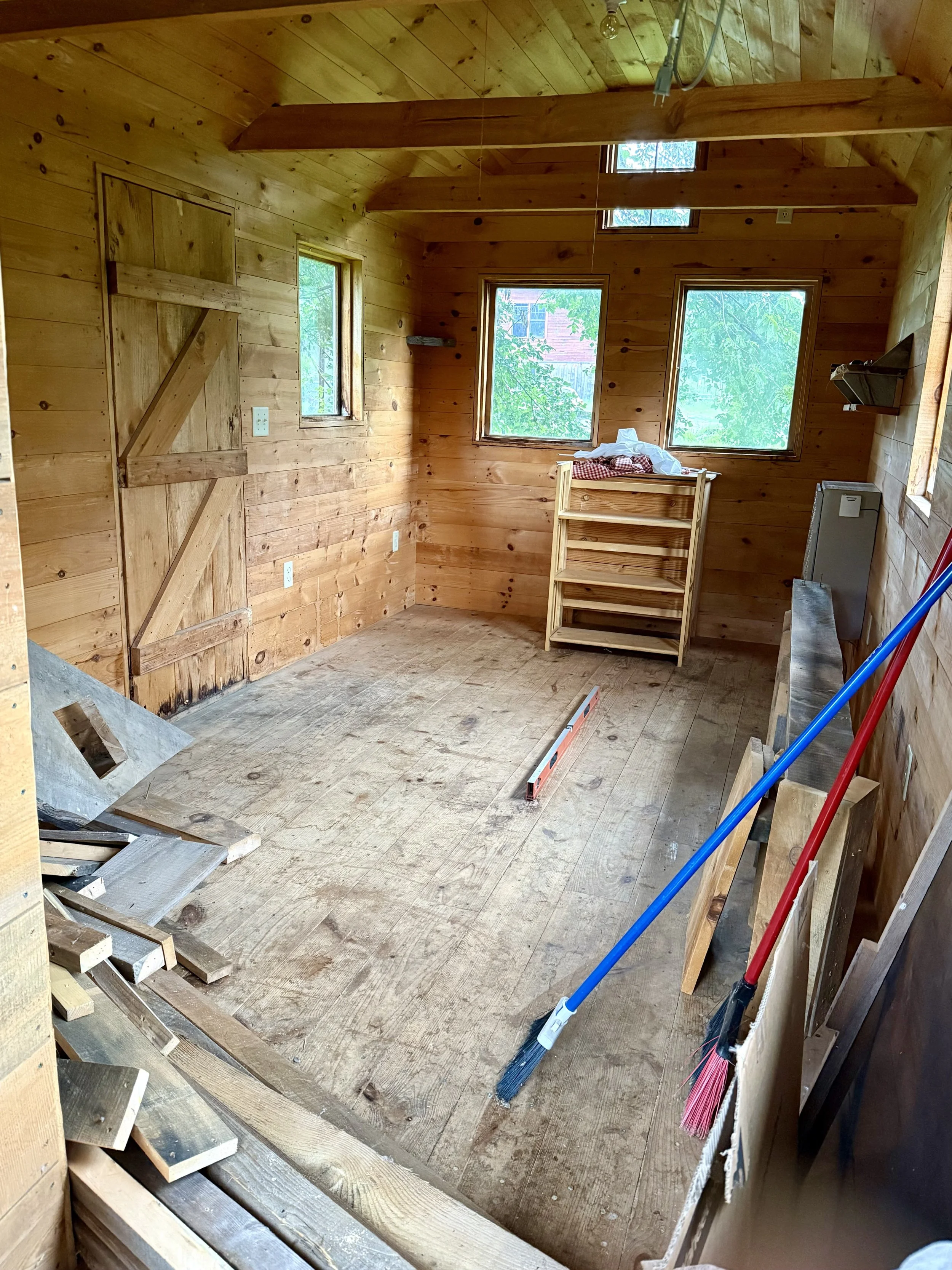 Interior of a partially renovated wooden room with unfinished floor, leaning tools, broom, and building materials, three windows letting in natural light, and a small shelving unit near the windows.