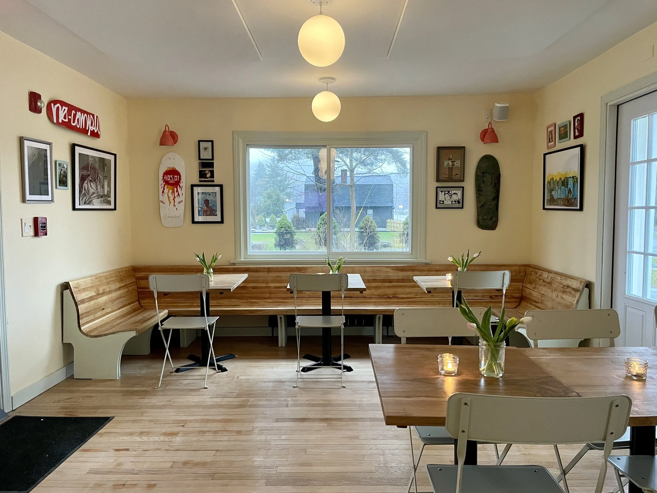 A cozy cafe interior with a corner booth, wooden tables, white chairs, potted tulips, candles on the tables, wall art, and large windows showing an outside view with trees.