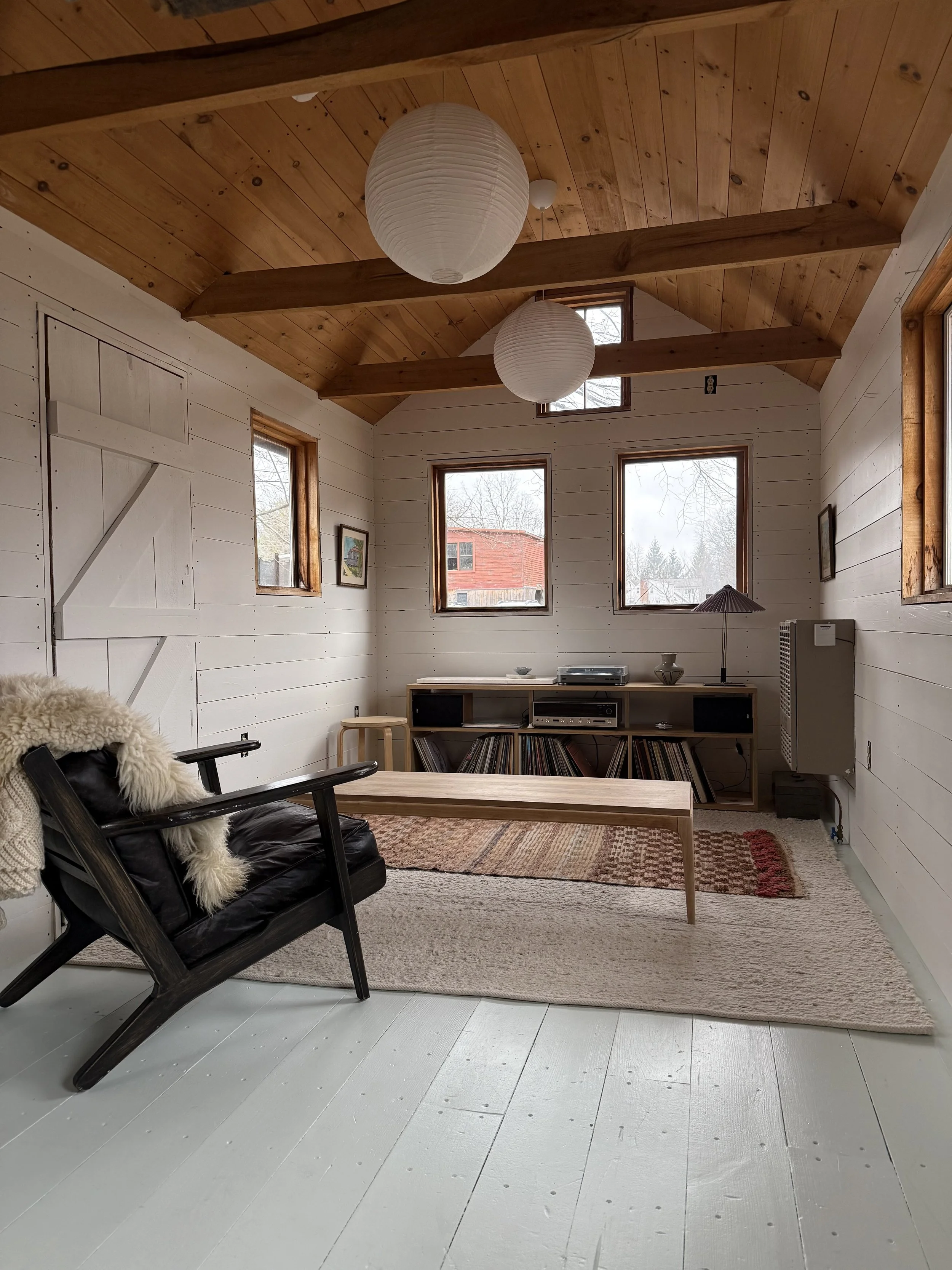 Cozy living room with white wooden walls and ceiling, featuring windows that let in natural light. Contains a black leather armchair with a fur blanket, a wooden coffee table, a media console with records, and two paper lantern pendant lights hanging