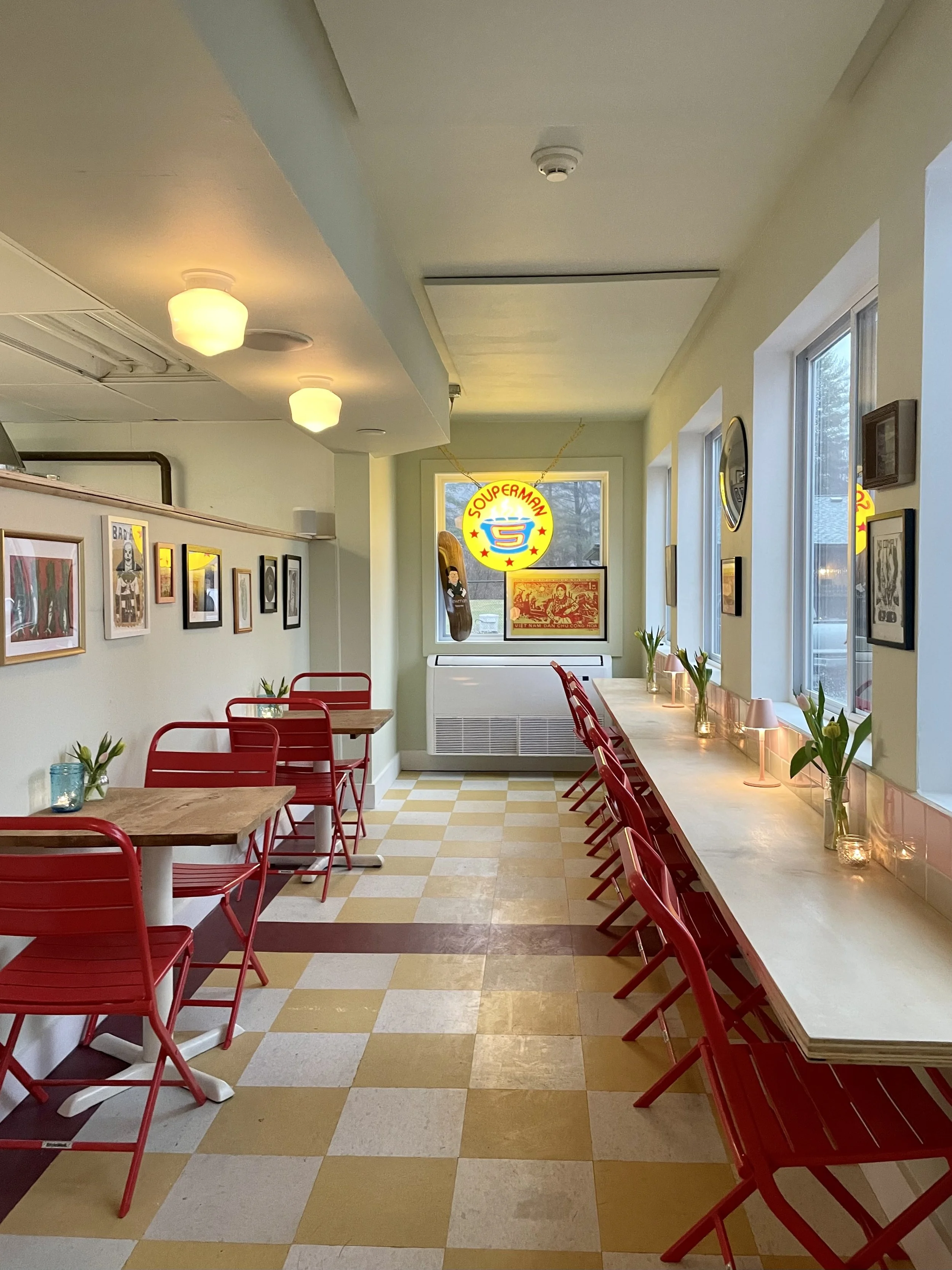Bright café interior with red chairs set at wooden tables, framed artwork on pale green walls, sunlight through large windows, and a circular sign that reads 'Souperman' in the background.