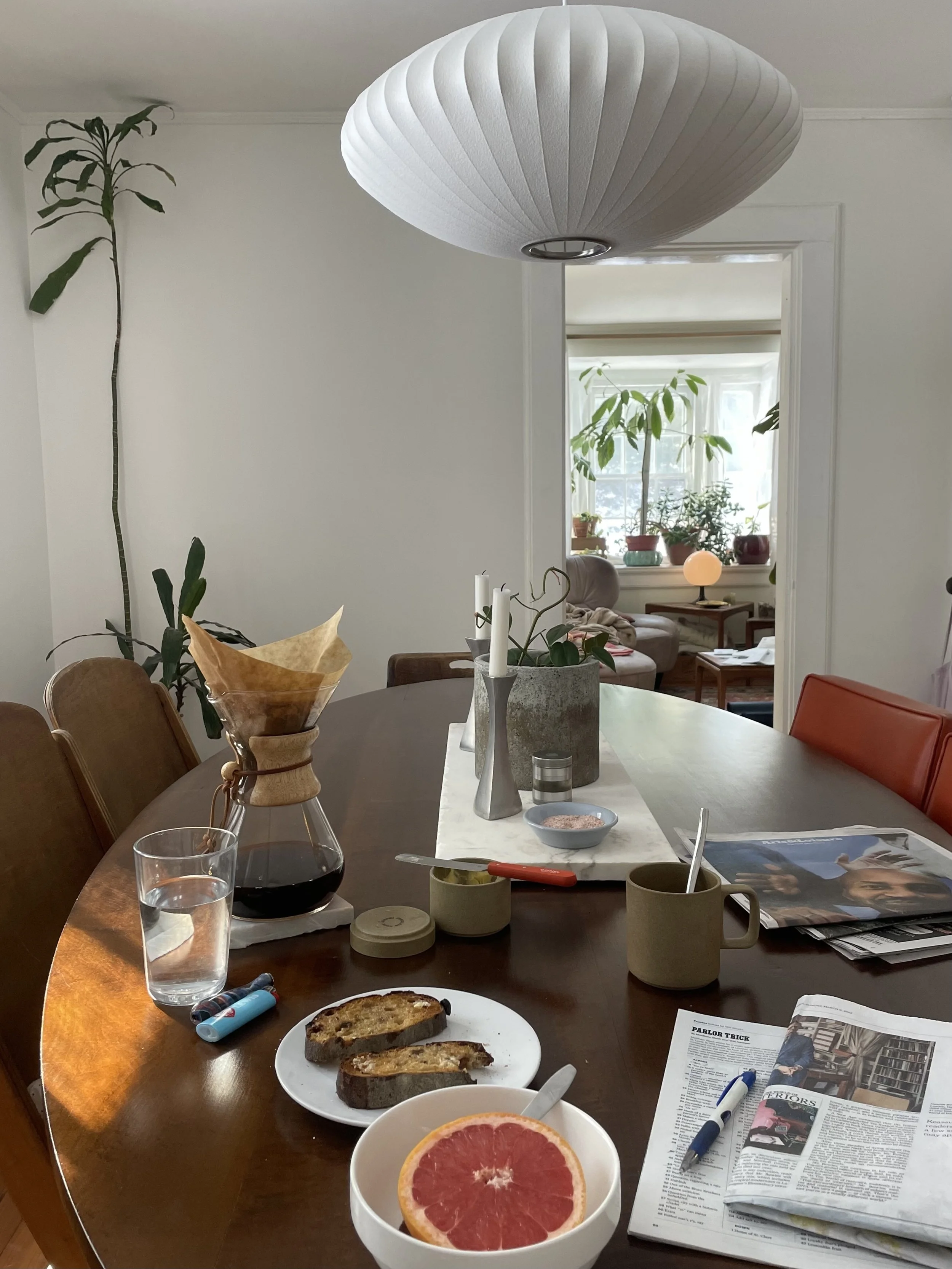 A dining room table with a white runner in the center, topped with two white candles, a potted plant, and kitchen items. On the table are three slices of toasted bread, a bowl with a grapefruit half, a glass of water, a coffee drip cone, a mug, a pen