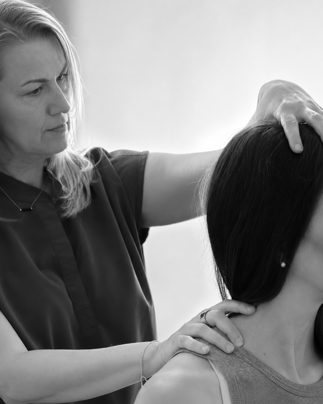 A woman with blonde hair examining a person's neck and shoulder, possibly a healthcare professional performing a medical or therapeutic assessment.