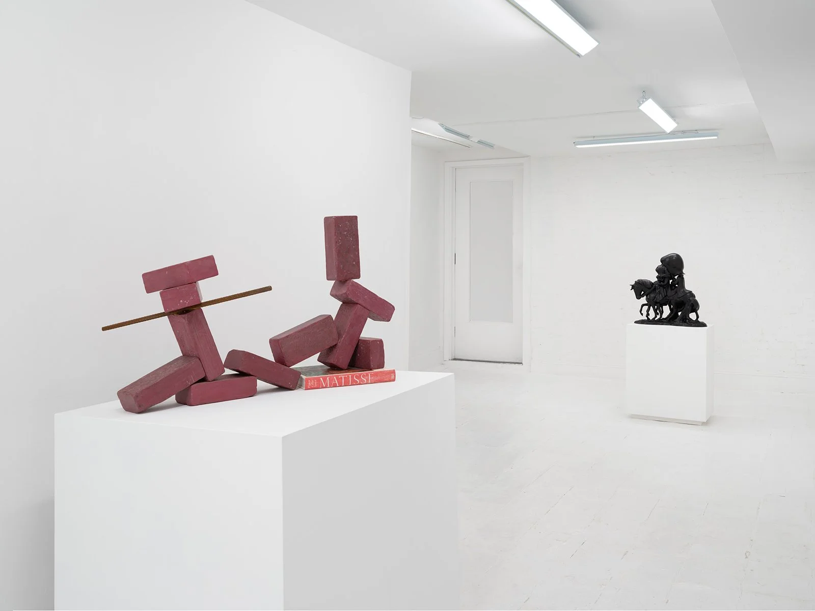 Art gallery with white walls, floor and ceiling, two sculptures displayed on white pedestals, fluorescent ceiling lights.