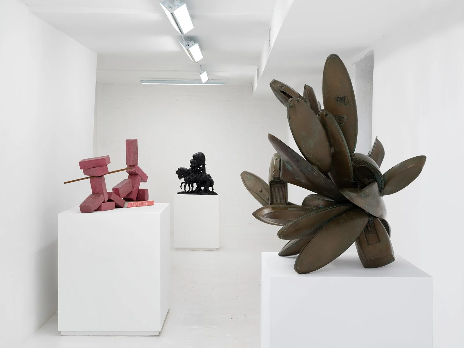 Art gallery with white walls, floor and ceiling, three sculptures displayed on white pedestals, fluorescent ceiling lights.