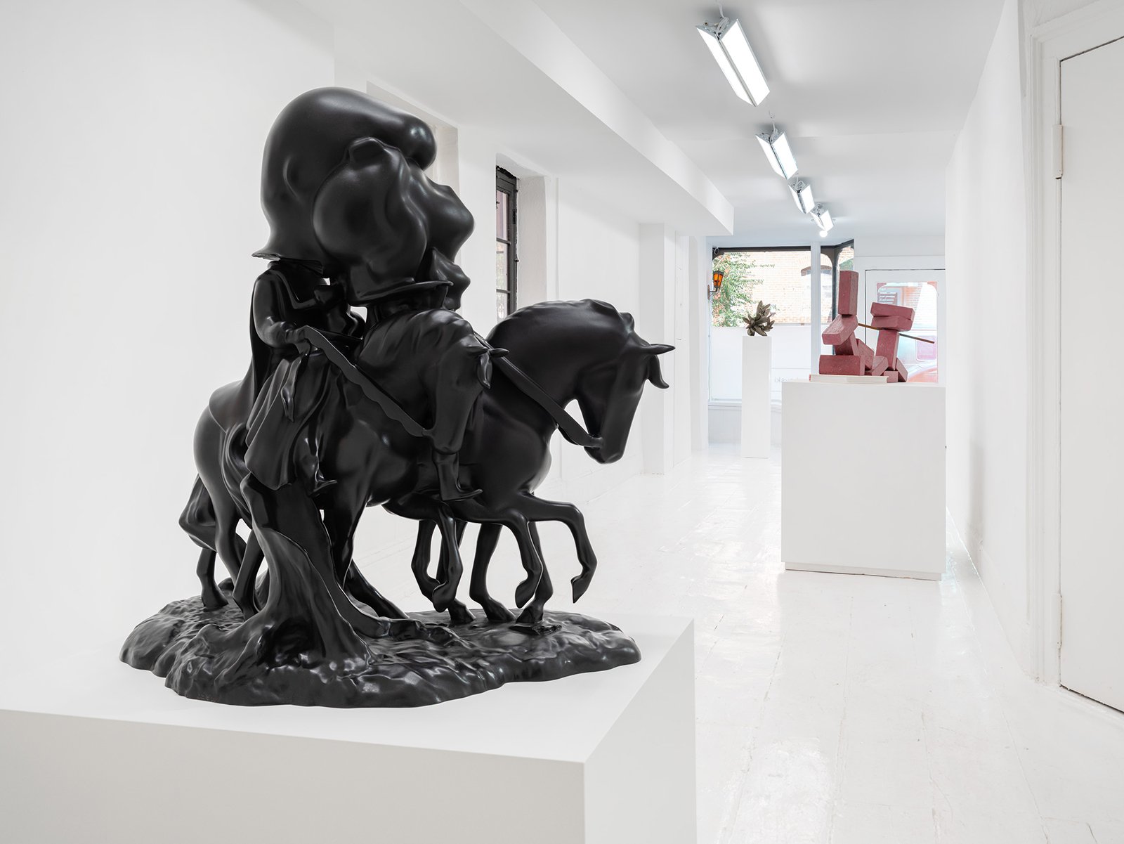Art gallery with white walls, floor and ceiling, three sculptures displayed on white pedestals, fluorescent ceiling lights and a window in the background.
