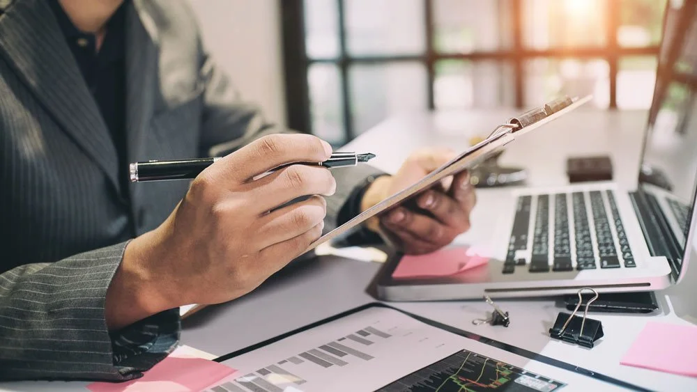 Person in business attire writing on a clipboard at a desk with a laptop, financial documents, and office supplies.