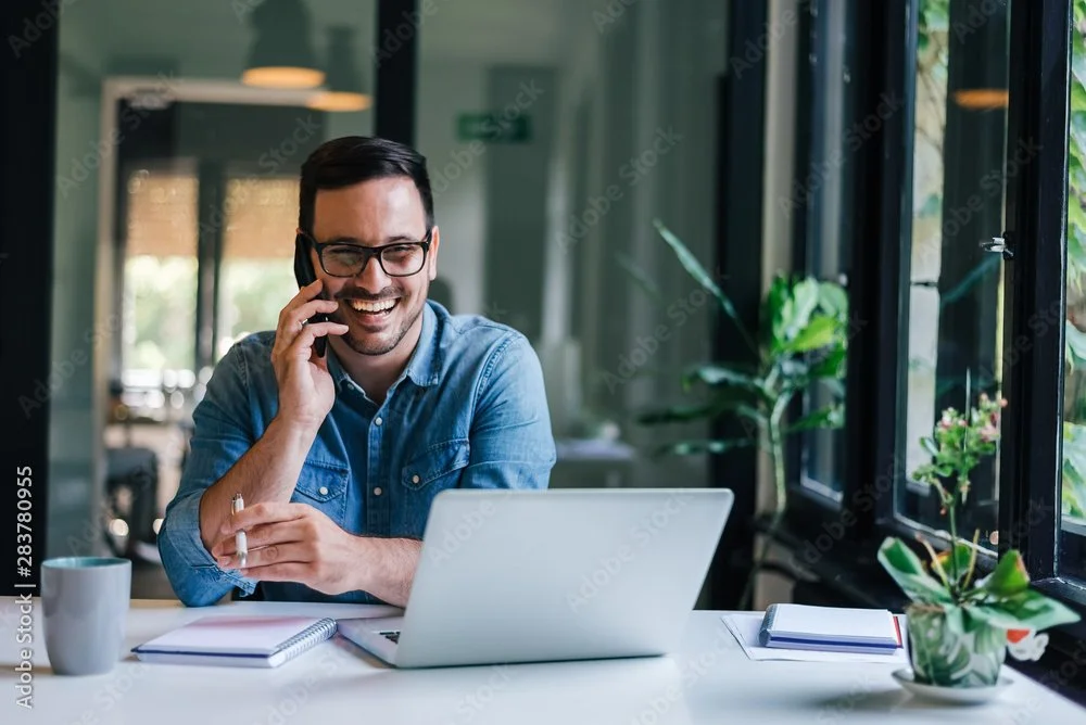 A man with dark hair and glasses smiling while talking on a cell phone at a desk with a laptop, notebooks, and a coffee mug in a bright, modern office or cafe with large windows and plants.
