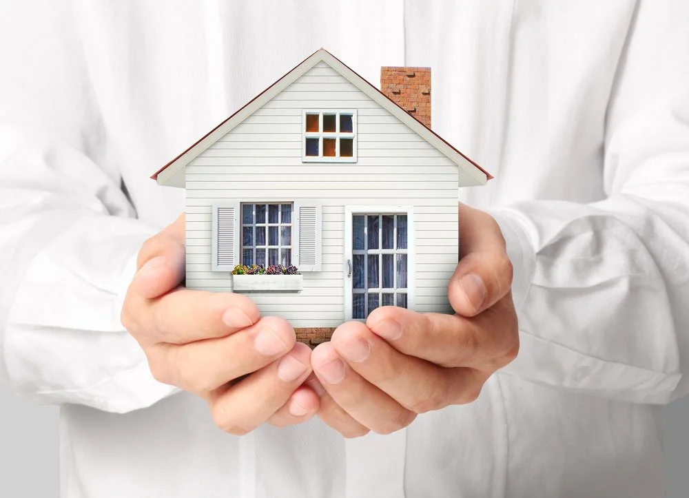Person holding a small model house with white siding, blue shutters, a flower box, and a chimney.
